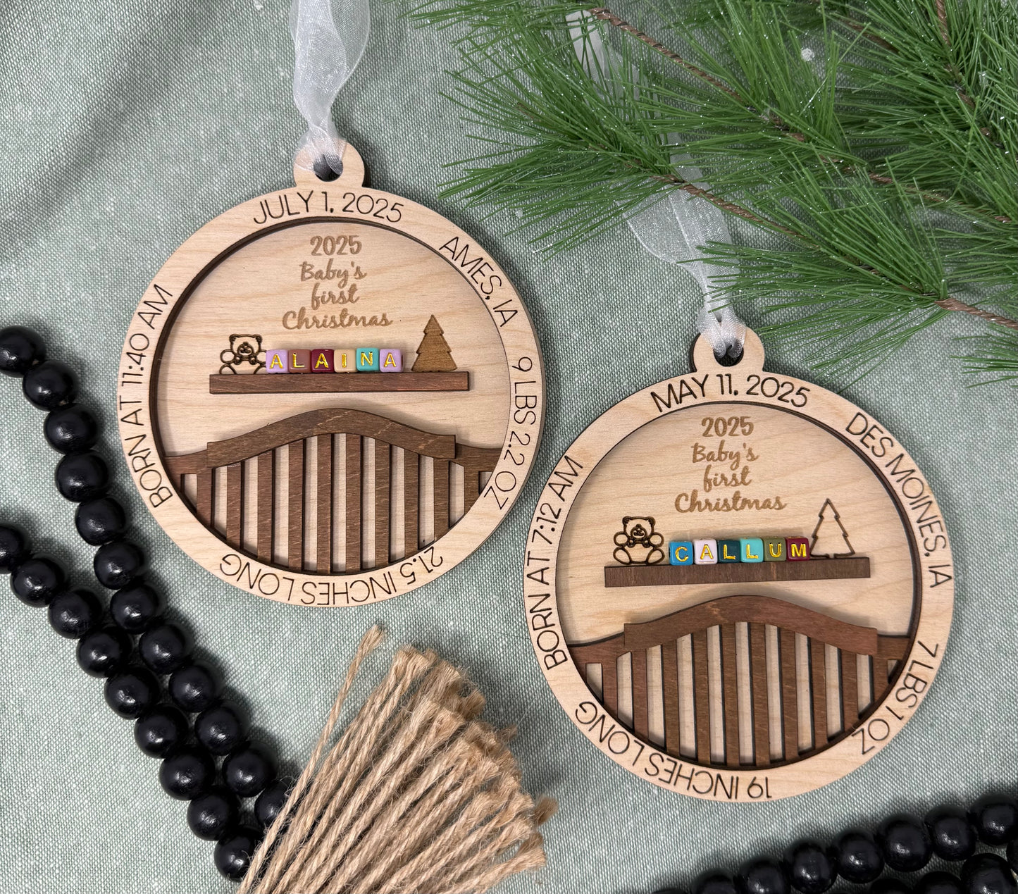 Two wooden Christmas ornaments with engraved text and designs on a gray surface with greenery.