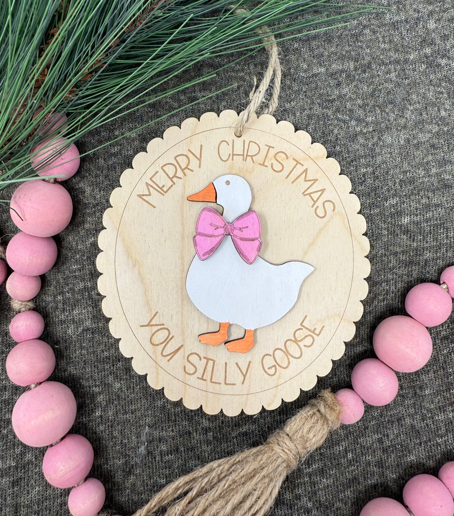 Wooden Christmas ornament with a goose design and text, surrounded by pink beads and greenery.