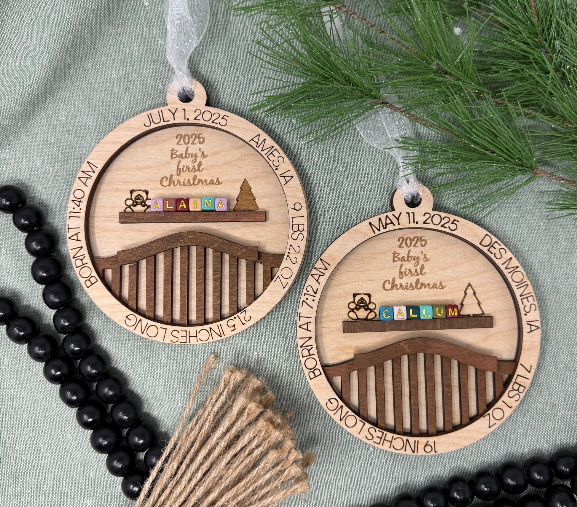Two wooden Christmas ornaments with engraved text and designs on a gray surface with greenery.