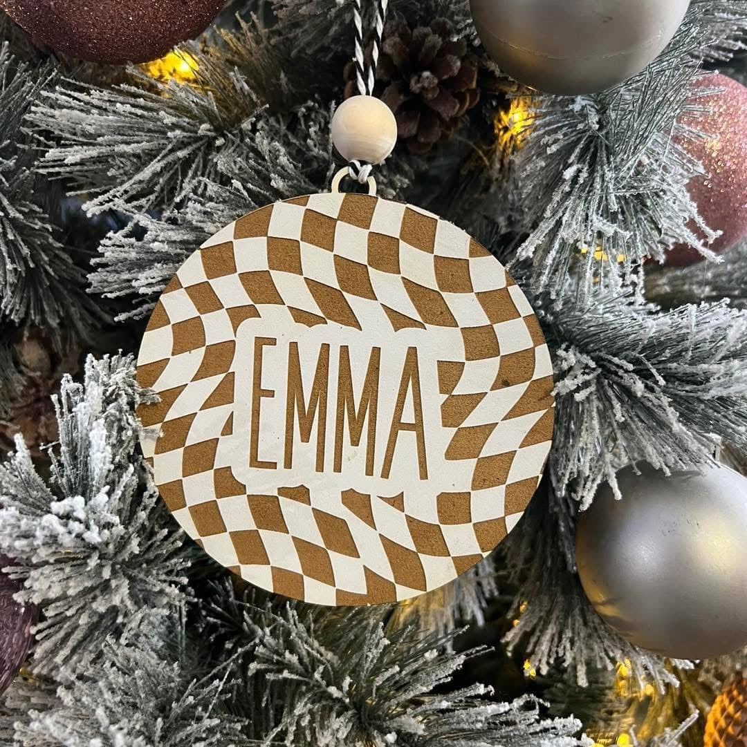 Decorative ornament with 'EMMA' on a checkered pattern, hanging on a Christmas tree.