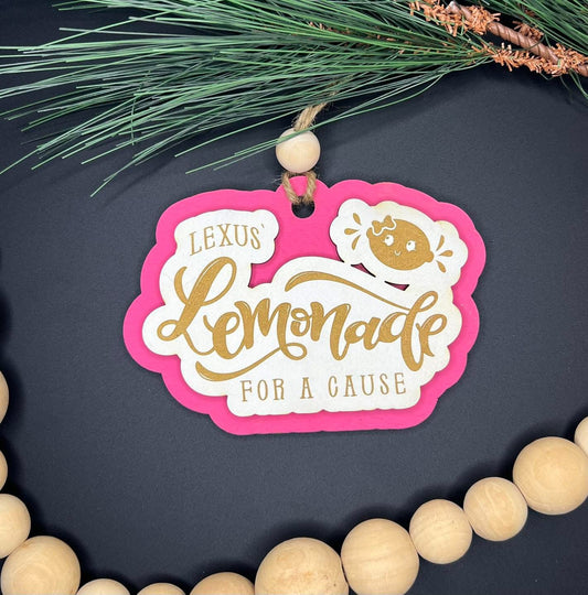 Decorative tag with 'Lexus Lemonade for a Cause' on a dark background with greenery and wooden beads.