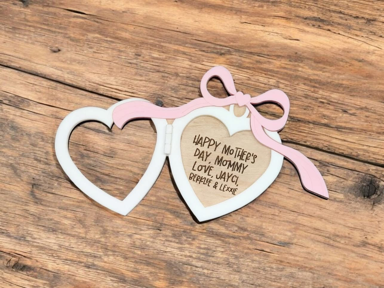 A heart-shaped, beige wooden magnet with a pink ribbon, and text that reads 'Happy Mother's Day' along with additional personalized text that says 'Love, [space for name], [space for name]' on a rustic wooden background.