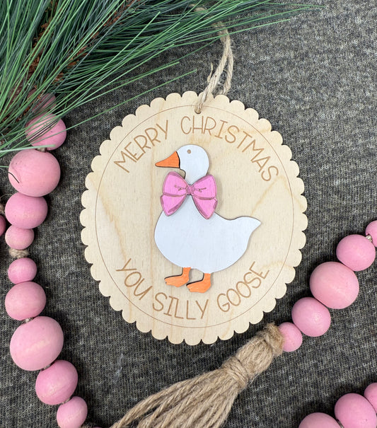 Wooden Christmas ornament with a goose design and text, surrounded by pink beads and greenery.