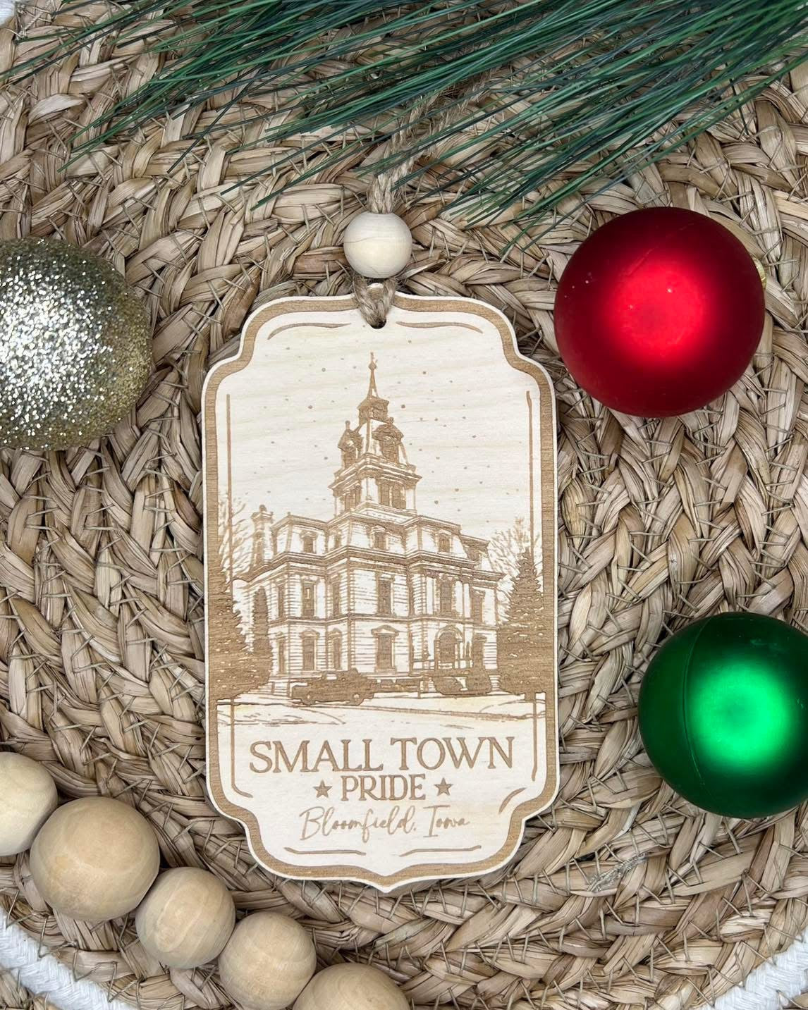 Decorative tag with 'Small Town Pride' text and building illustration, surrounded by Christmas ornaments on a woven mat.